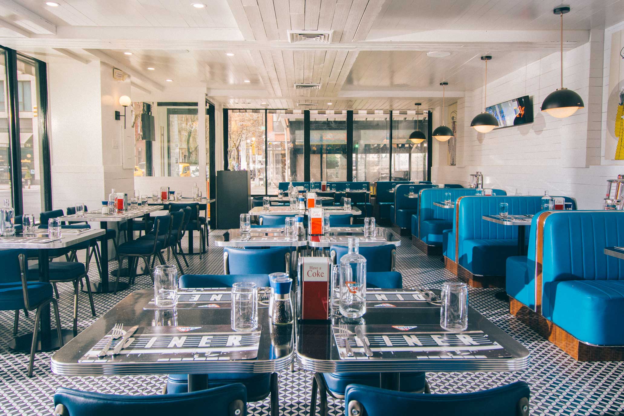 The bright and spacious dining area of Diner 24 NYC, featuring modern retro-inspired blue booths, sleek black-and-white tiled floors, and sunlit interiors through large windows. Known for its reputation as one of the top-rated diners in NYC, this cozy yet stylish space perfectly complements its menu of high-quality diner food, NYC’s favorite comfort food, and options catering to vegetarians and vegans. A destination for the best burgers near me in NYC and late-night dining in Manhattan, Diner 24 NYC delivers an exceptional 24/7 dining experience.