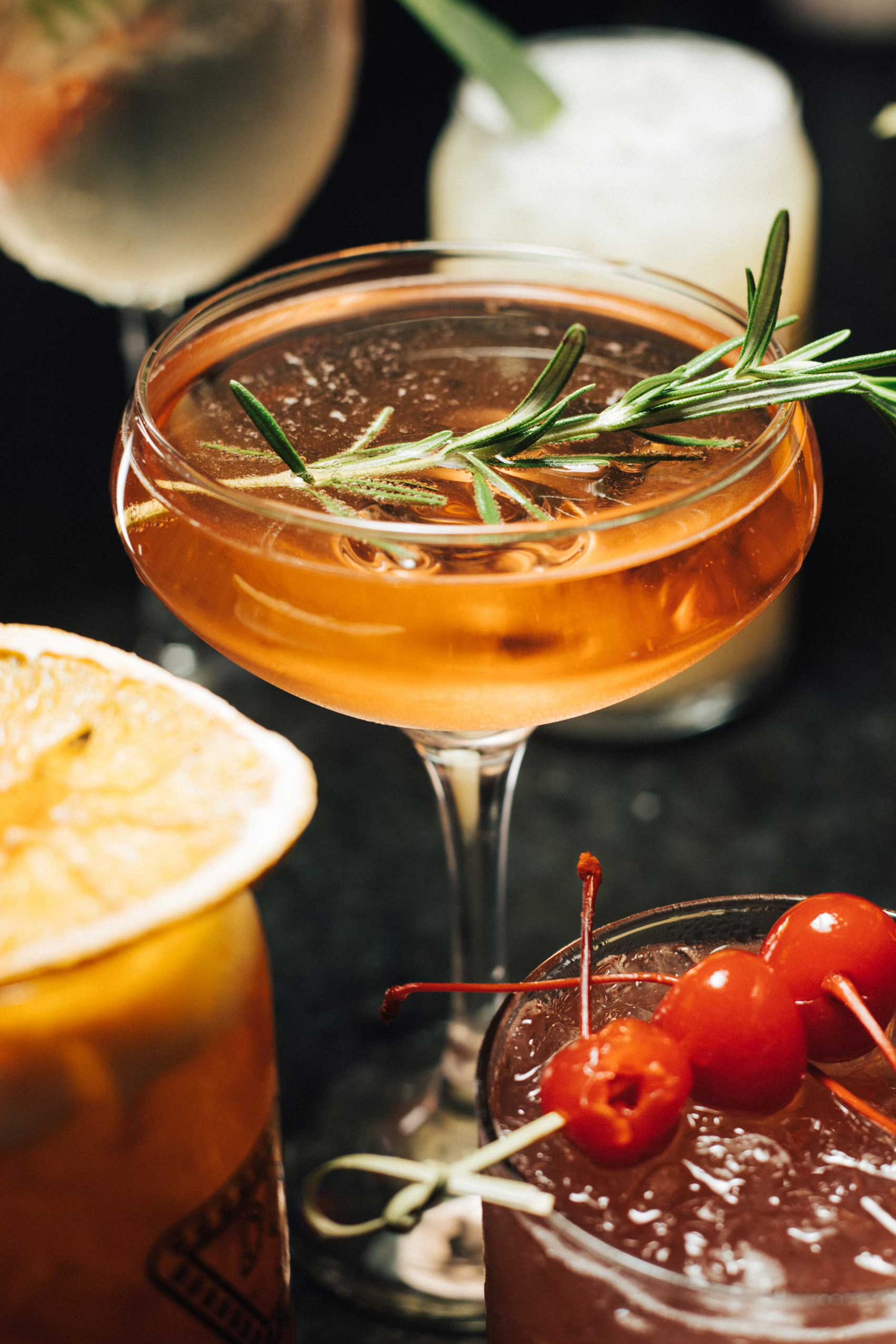 A sophisticated Gotham Sour cocktail elegantly presented in a coupe glass, showcasing its amber hue and topped with a fresh rosemary sprig garnish. The cocktail is surrounded by other vibrant drinks, including one with glistening maraschino cherries and a citrus-infused glass, creating a captivating visual of NYC’s dynamic mixology scene. Perfect for savoring in NYC's top-rated diners or pairing with high-quality diner food at late-night spots in Manhattan. This Gotham Sour is a refreshing take on classic cocktails, ideal for 24/7 diners in NYC.