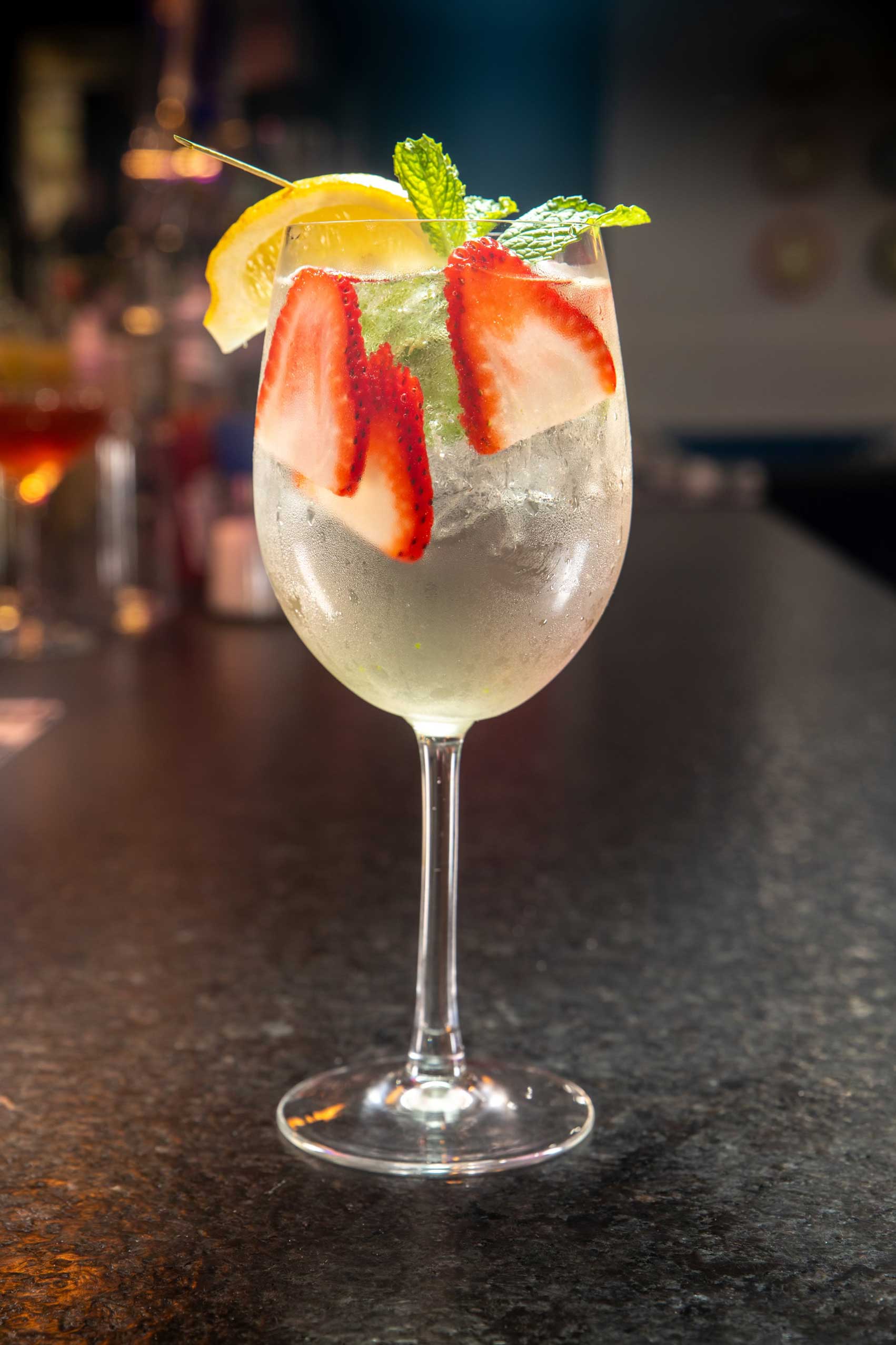 A refreshing Gramercy-Garden-Fizz cocktail served in a crystal-clear wine glass, filled with ice cubes and garnished with fresh strawberry slices, a sprig of mint, and a wedge of lemon. The translucent drink sparkles under soft lighting, showcasing its vibrant ingredients that blend citrusy tang with subtle berry sweetness and a hint of herbal mint. Perfect for savoring in NYC’s top-rated diners or enjoying as a sophisticated addition to late-night dining in Manhattan. This cocktail pairs beautifully with high-quality diner food and NYC's favorite comfort dishes.