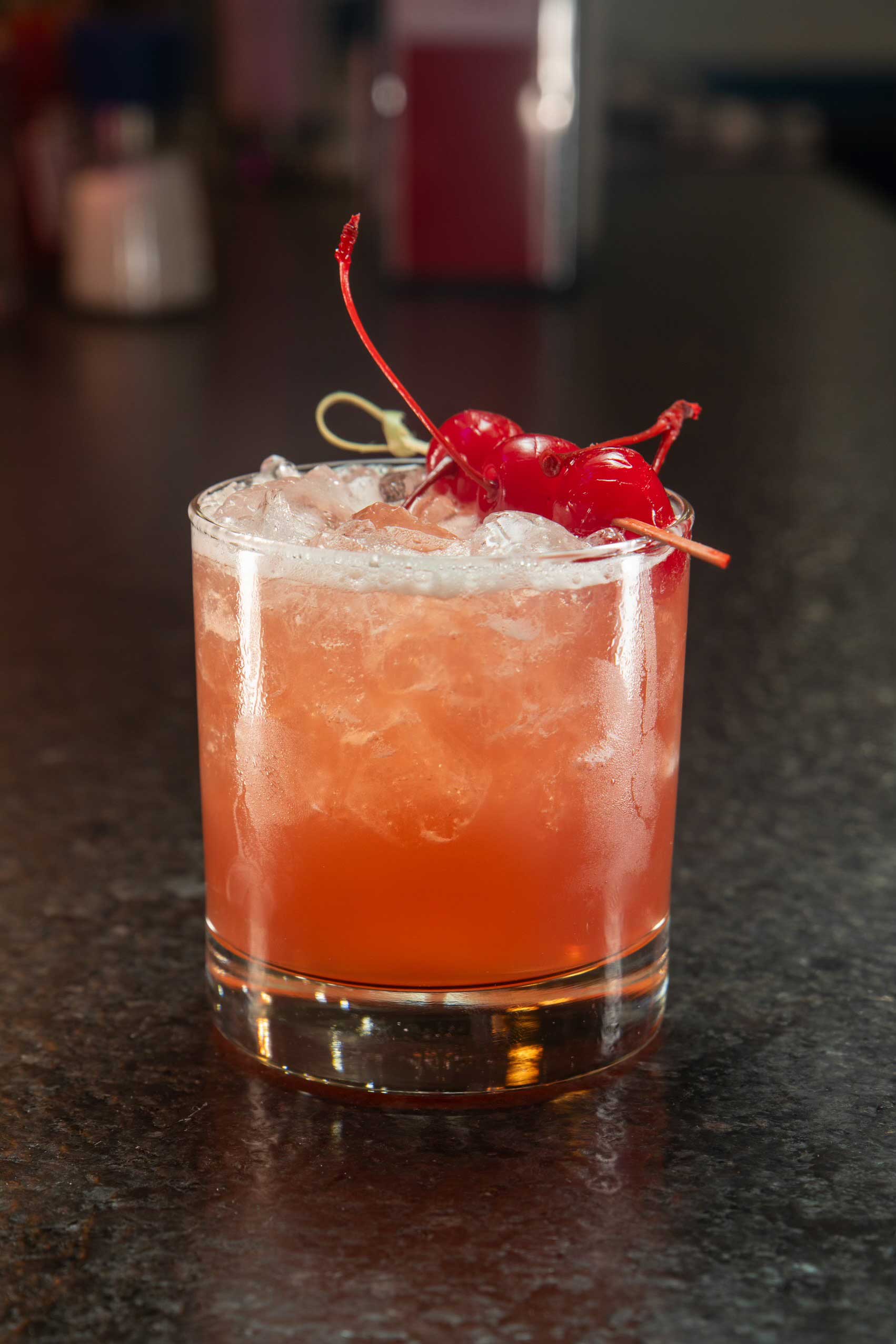 A refreshing Lavender-Manhattan cocktail served in a short tumbler glass, filled with crushed ice and garnished with vibrant maraschino cherries. This cocktail features a delicate blend of lavender-infused flavors with the classic warmth of Manhattan's signature whiskey and vermouth base. The bright cherry garnish adds a pop of color against the soft orange-pink hues of the drink, creating an inviting presentation. Perfectly crafted to complement NYC’s favorite comfort food and elevate any dining experience at one of the top-rated diners in NYC.