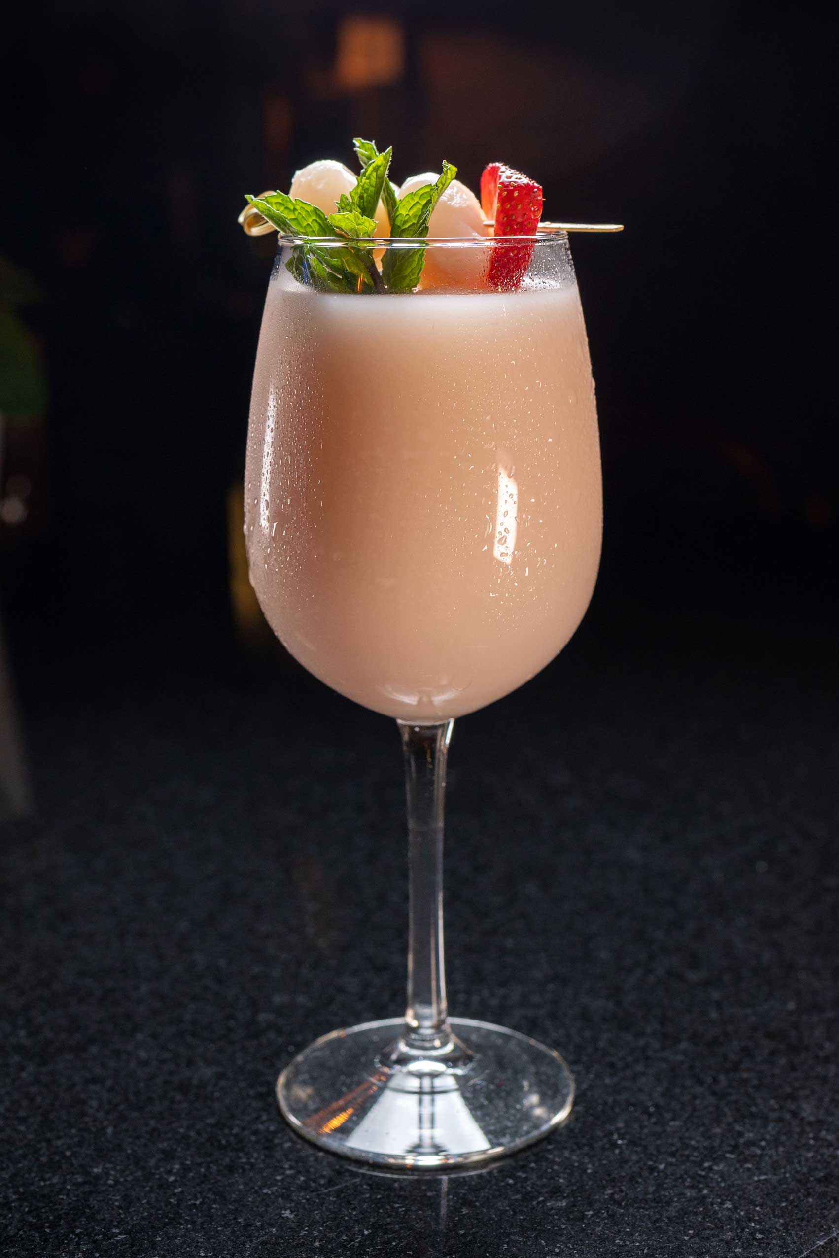 A luxurious Lychee-Coconut-Frosé cocktail elegantly served in a tall wine glass, blending creamy coconut and refreshing lychee flavors with a rosé wine base. Garnished with fresh mint leaves, lychee fruit, and a slice of strawberry, this tropical-inspired drink exudes sophistication. The light blush hue contrasts beautifully against the dark background, while the condensation on the glass highlights its refreshing appeal. This cocktail is a standout offering at NYC’s top-rated diners, perfect for pairing with high-quality diner food or enjoying as a standalone treat during a late-night dining experience in Manhattan.