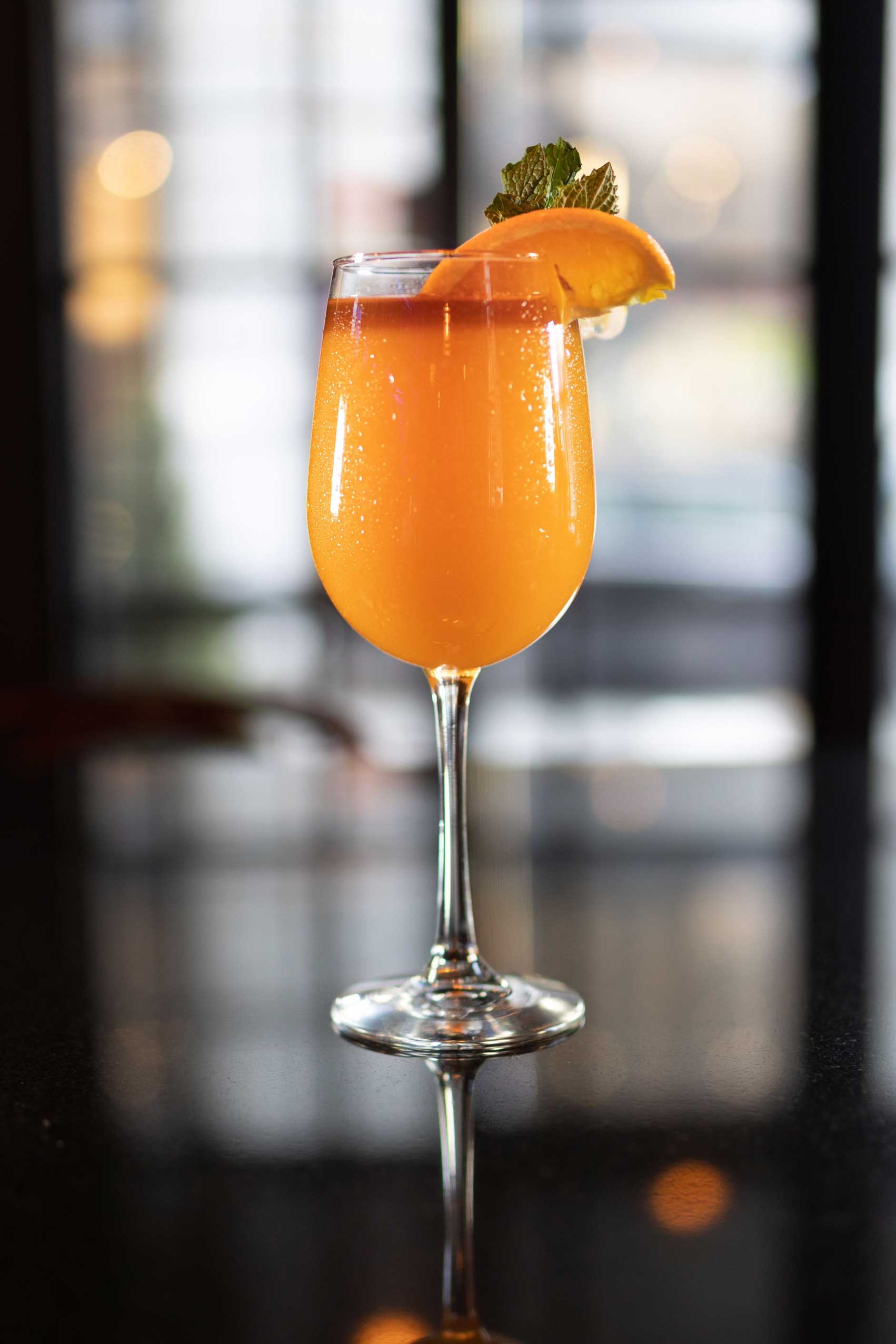 A refreshing orange cocktail served in a stylish wine glass, garnished with a slice of fresh orange and a sprig of mint. The drink's vibrant color glows against the sleek, reflective bar surface, while a softly lit background adds an elegant touch. This visually appealing beverage is ideal for enjoying at one of NYC's top-rated diners, known for their high-quality diner food and creative drink offerings. Perfect for a casual brunch or an evening out, this cocktail complements both vegetarian-friendly and vegan-friendly breakfast options, as well as late-night dining in Manhattan.