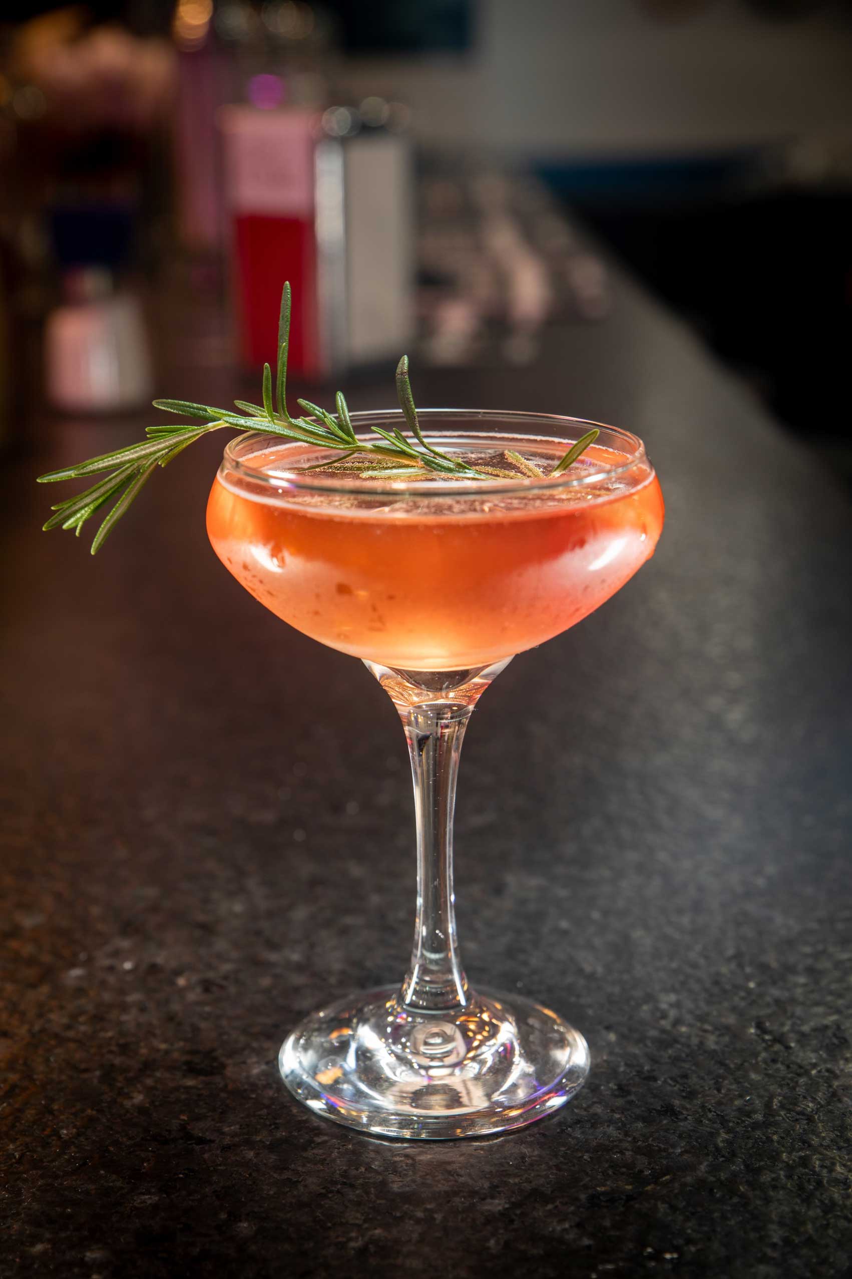 Elegant cocktail served in a sophisticated coupe glass at one of NYC's top-rated diners. This drink features a light amber hue with a sprig of fresh rosemary delicately placed on top, exuding a refined and aromatic touch. Set against the sleek, polished bar surface, this cocktail offers a perfect combination of flavor and presentation. Ideal for enjoying during a late-night dining experience in Manhattan or as a highlight of your visit to one of the best 24/7 diners in NYC, this drink embodies the creativity and quality of high-end diner beverages.