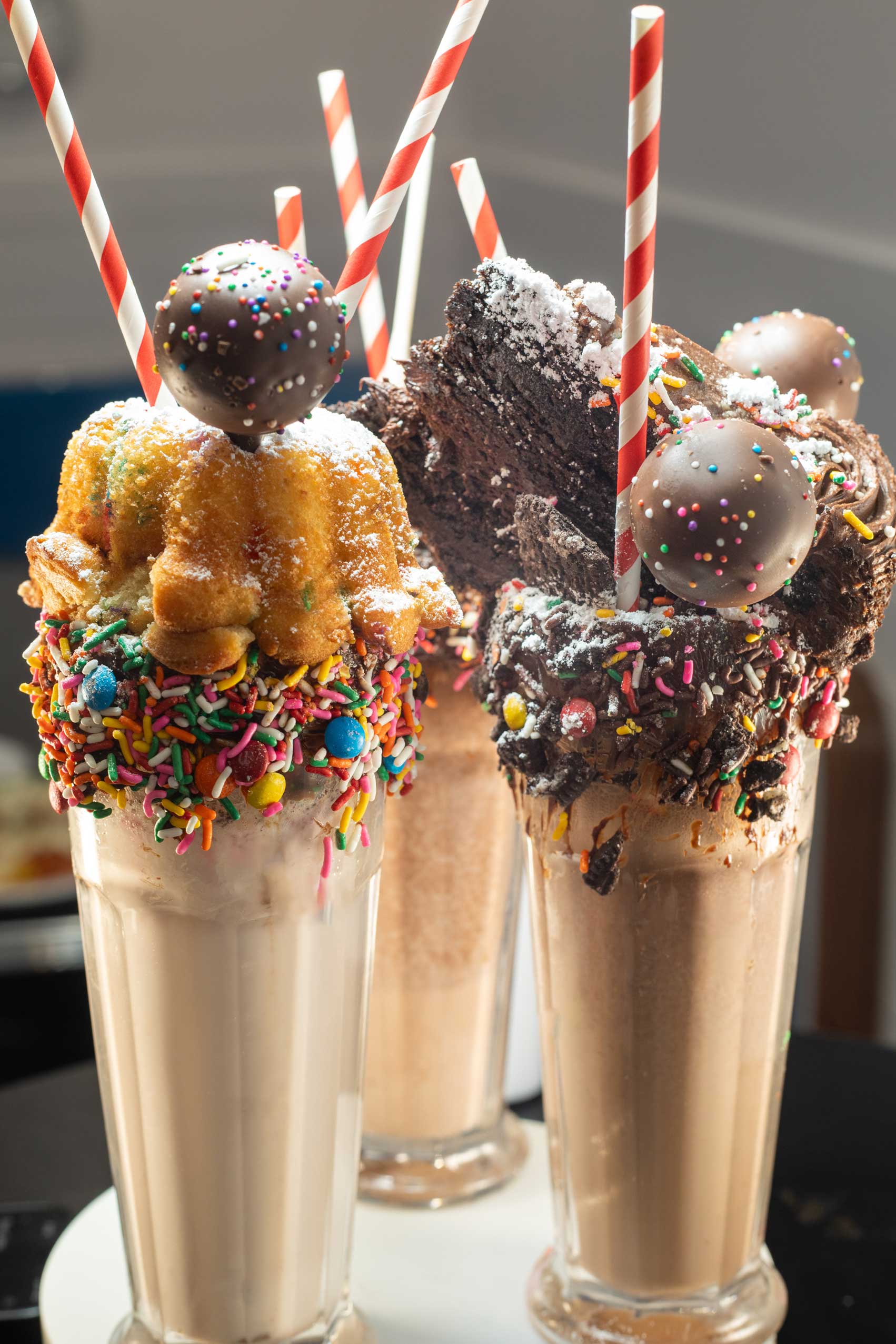 A trio of indulgent milkshakes served in tall glasses, each topped with a decadent combination of sweets. One milkshake is crowned with a sprinkle-covered bundt cake, another with rich chocolate brownies, and the third with chocolate truffles. The rims of the glasses are coated with vibrant rainbow sprinkles, candy pieces, and chocolate drizzle, adding a whimsical touch to these over-the-top desserts. These extravagant milkshakes capture the essence of comfort food, blending creativity and indulgence, making them a perfect treat at Diner 24 NYC, a top-rated diner for high-quality American comfort food in NYC.